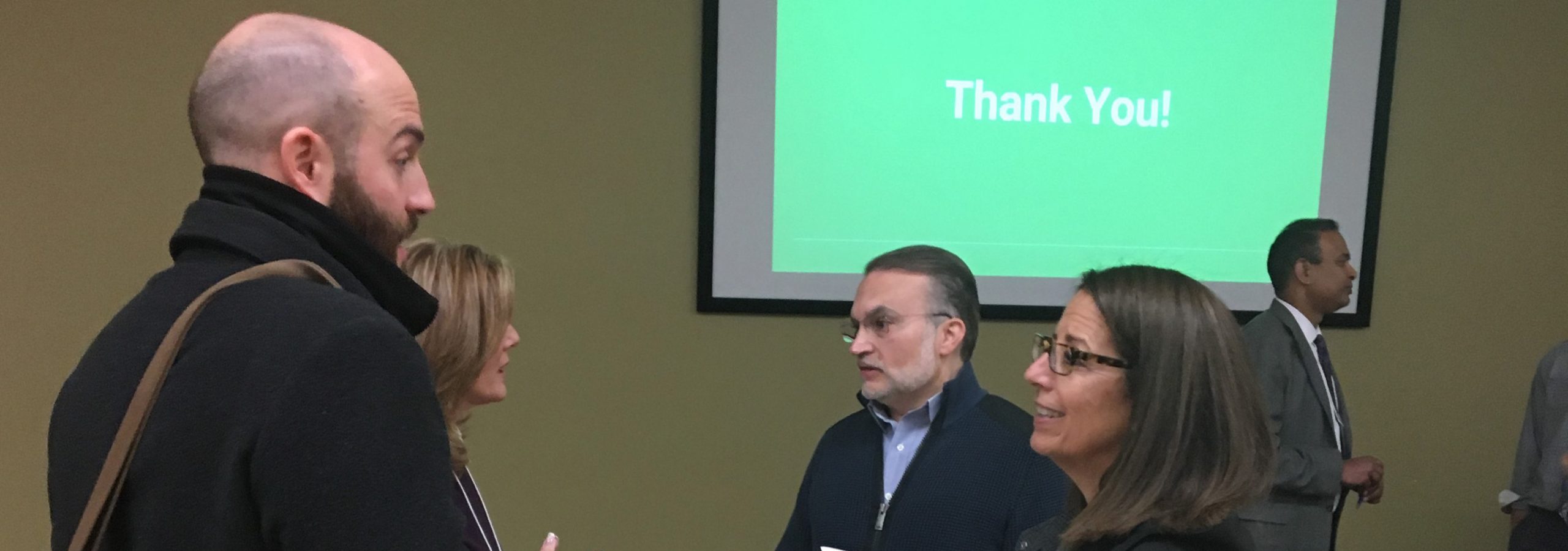 Photo of two men and two women talking with the end of a Project Advisory Committee PowerPoint presentation in background